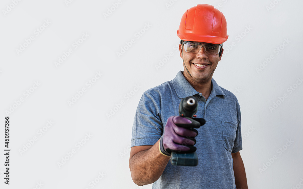 Confident young Latin male carpenter in hardhat holding drill and looking at camera with smile while standing against white background. Always ready to help. Confident young Latin male carpenter in hardhat holding drill and looking at camera with smile while standing against white background. Always ready to help.