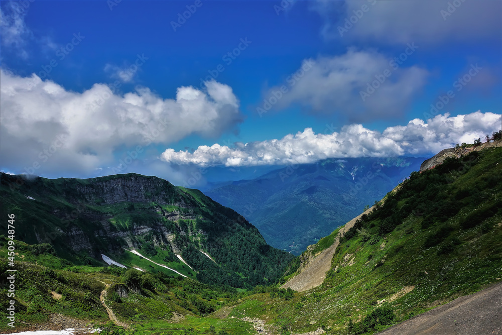Green grass and wildflowers grow on the mountain slopes. Patches of melted snow are visible among the greenery. There are picturesque cumulus clouds in the blue sky. A sunny summer day. Caucasus