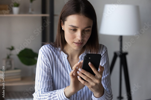 Young Caucasian woman hold use modern cellphone text message online on modern gadget. Focused millennial female look at smartphone screen talk speak on smartphone video webcam call.