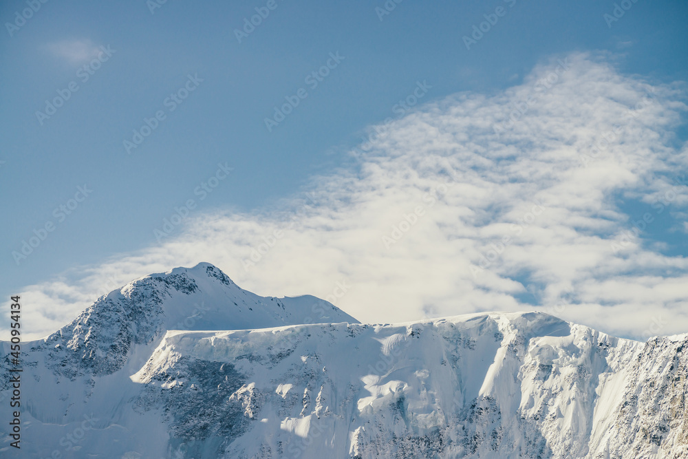 Minimalist alpine landscape with high snow-covered mountain with snowy peaked top under cirrus clouds. Beautiful mountain scenery with great snow-white sharp pinnacle in cloudy sky. Awesome mountains.