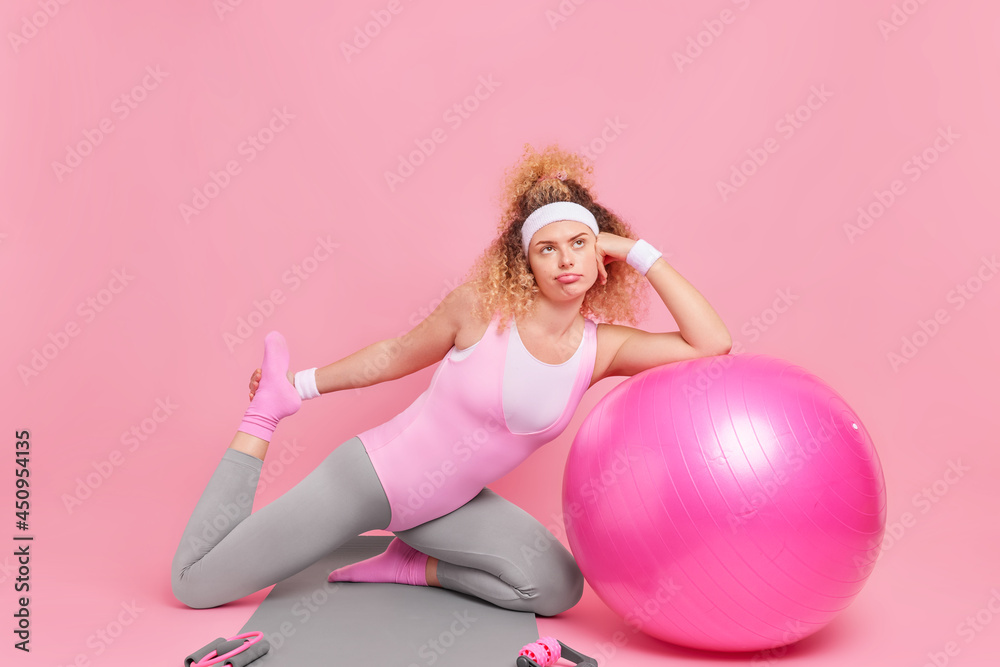 Bored unmotivated woman with curly hiar doesnt want to do gymanstics stretches legs on fitness mat leans on inflated swiss ball dressed in bodysuit isolated over pink background. Workout and people Bored unmotivated woman with curly hiar doesnt want to do gymanstics stretches legs on fitness mat leans on inflated swiss ball dressed in bodysuit isolated over pink background. Workout and people