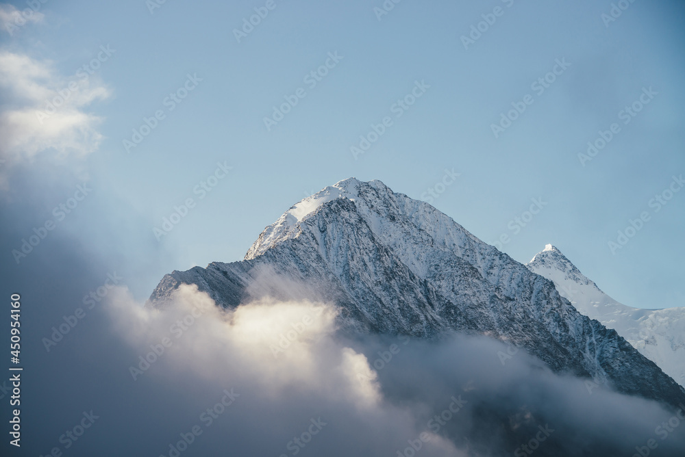 Beautiful view of snow-capped mountains above thick clouds in sunshine. Scenic bright mountain landscape with white-snow peak among dense low clouds in blue sky. Wonderful scenery with snowy pinnacle.
