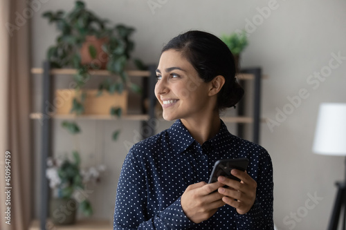 Smiling millennial Indian woman use smartphone look in window distance dreaming or thinking. Happy young biracial female speak on video webcam call on cellphone make plans or visualize.