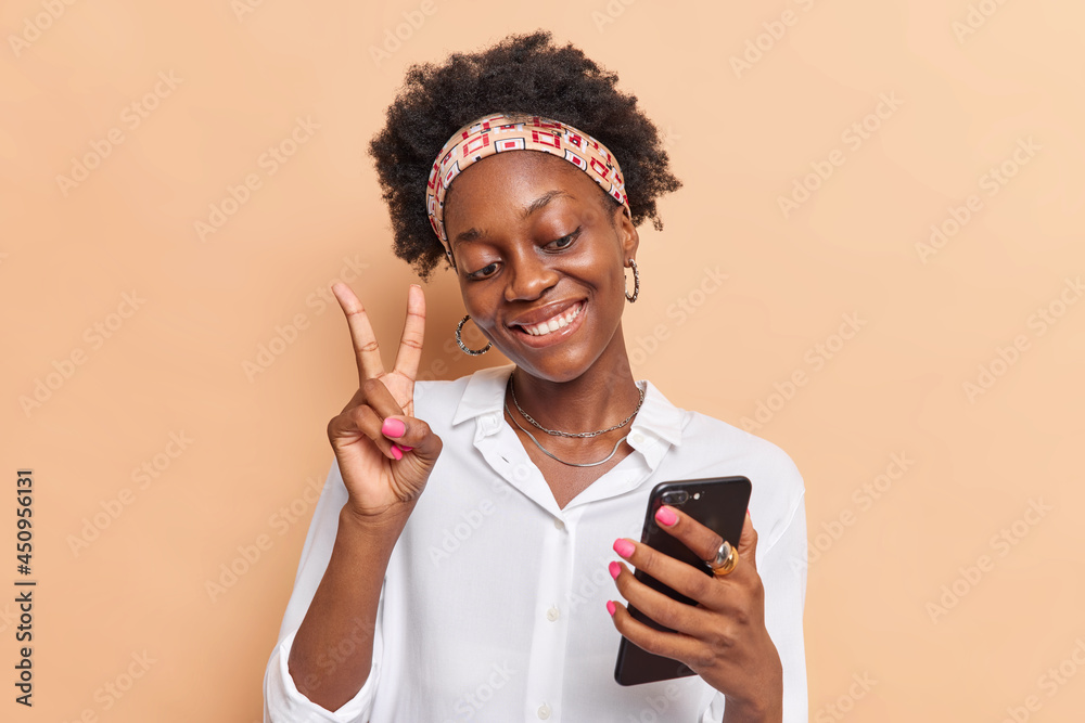 Happy teenage girl with dark skin curly Afro hair makes peace gesture takes selfie or makes video call via smartphone wears headband white shirt isolated over beige bacground. Technology concept Happy teenage girl with dark skin curly Afro hair makes peace gesture takes selfie or makes video call via smartphone wears headband white shirt isolated over beige bacground. Technology concept