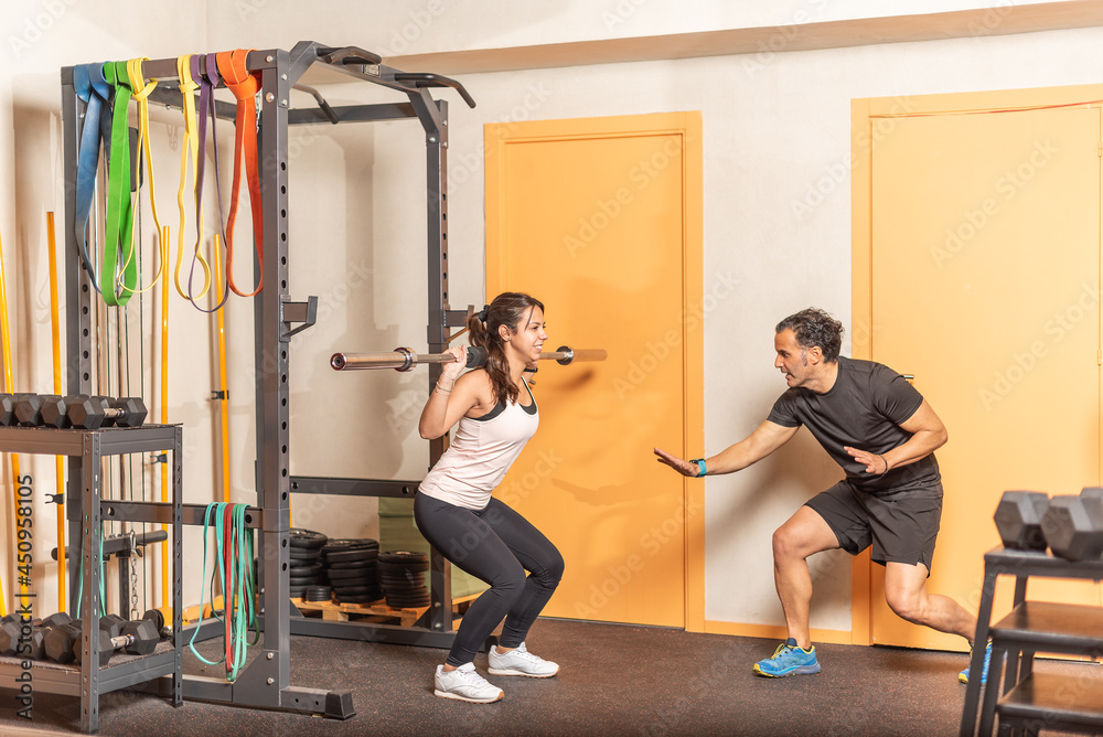 Fototapeta premium Sportswoman doing squats with bar with trainer in gym