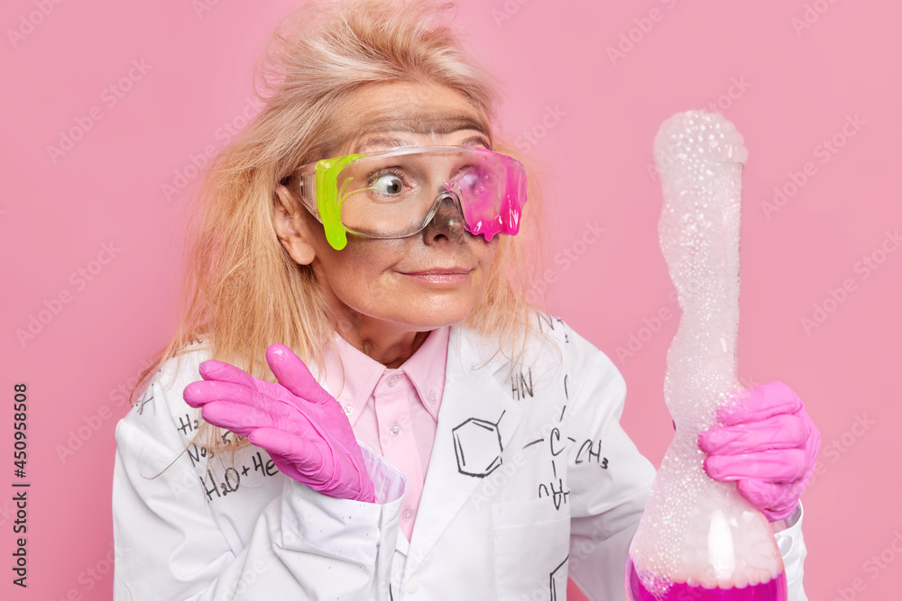 Middle aged female scientific professor looks at explosion after mixing reagents in flask wears protective glasses white coat surprised with result of experiment works in laboratory. Chemistry concept Middle aged female scientific professor looks at explosion after mixing reagents in flask wears protective glasses white coat surprised with result of experiment works in laboratory. Chemistry concept