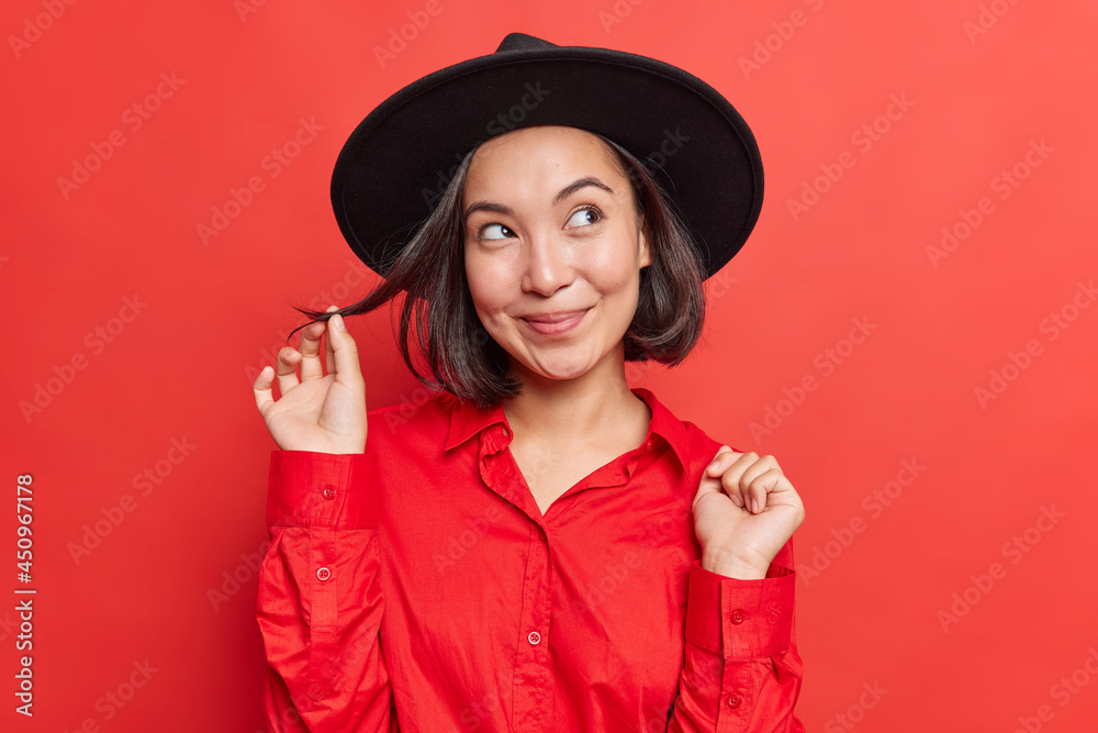 Attractive brunette young Asian woman without makeup with dreamy expression thinks about something wonderful curls hair wears black hat red shirt poses indoor against vivid studio background Attractive brunette young Asian woman without makeup with dreamy expression thinks about something wonderful curls hair wears black hat red shirt poses indoor against vivid studio background