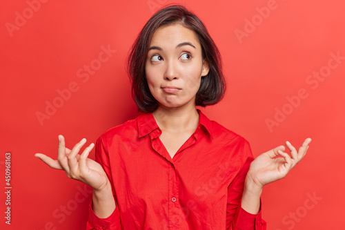 Clueless hesitant beautiful Asian woman with short dark hair spreads palms sideways shrugs shoulders has indecisive expression dressed in shirt stands against vivid red background. Who knows.
