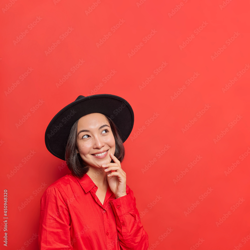 Vertical shot of thoughtful young Asian woman has glad face expression keeps hand on face focused above dressed in fashionable outfit poses against vivid red background copy space for your promo Vertical shot of thoughtful young Asian woman has glad face expression keeps hand on face focused above dressed in fashionable outfit poses against vivid red background copy space for your promo