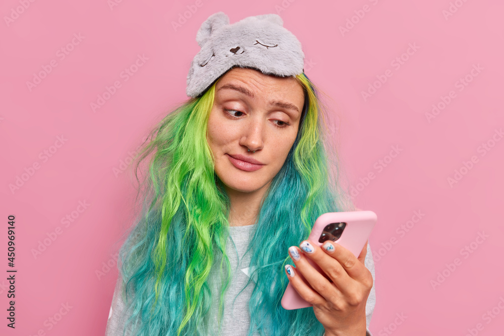 Upset hipster girl reads information on smartphone looks with unhappy expression gets message from formal boyfriend wears sleepmask on forehead isolated over pink background. Technology concept Upset hipster girl reads information on smartphone looks with unhappy expression gets message from formal boyfriend wears sleepmask on forehead isolated over pink background. Technology concept