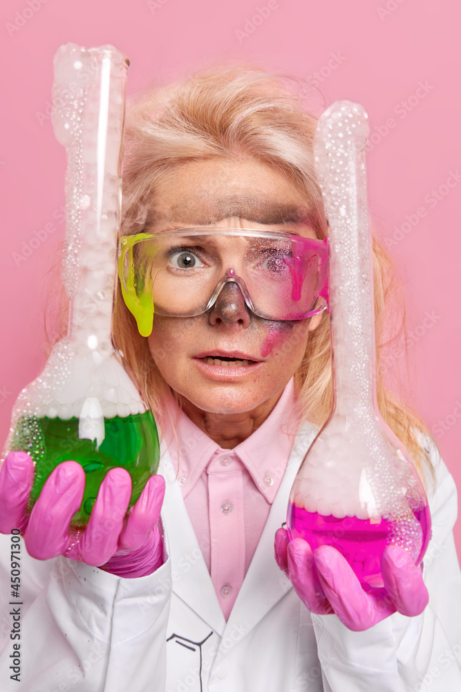 Puzzled shocked female chemist tests product samples mixes ingredients in flasks surprised with reagent reaction wears safety glasses white coat rubber gloves. Bubbles after solution distillation