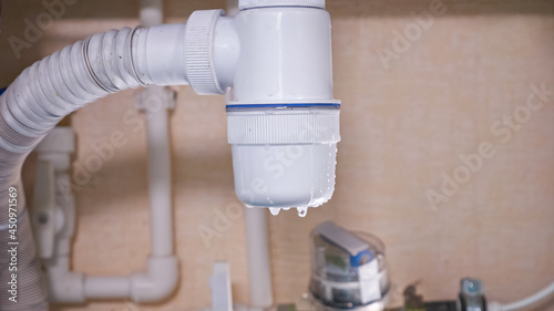 Dirty water drips down from damaged white plastic plumbing trap under sink in contemporary kitchen at bright light extreme close view