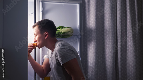 Careful young man opens door of modern fridge and eats delicious creamy eclair in secret turning back in dark empty kitchen at night