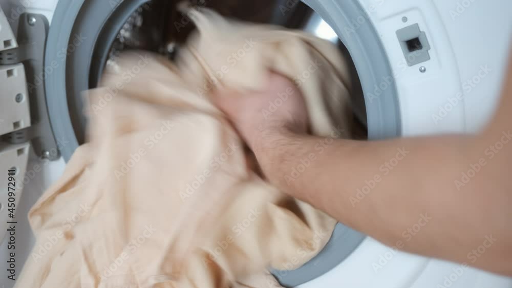 Man's hands loading the washing machine with beige bed linen, laundry at home. Man is doing his laundry and closing the door of washing machine. Everyday life, housework of young man. Man's hands loading the washing machine with beige bed linen, laundry at home. Man is doing his laundry and closing the door of washing machine. Everyday life, housework of young man.