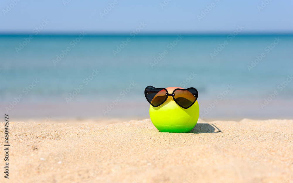 A colored ball with wearing heart-shaped sunglasses lies on a sandy beach against the backdrop of the sea.