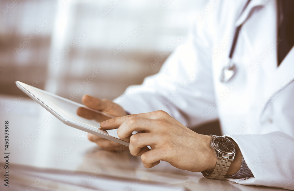 Fototapeta premium Unknown male doctor sitting and working with tablet computer in clinic at his working place, close-up. Young physician at work. Perfect medical service, medicine concept