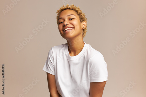Beautiful mixed-race female model with culry blonde short haircut, wearing nose ring, posing in white T-shirt with carefree, cheerful and happy face expression, isolated on pale pink bakground