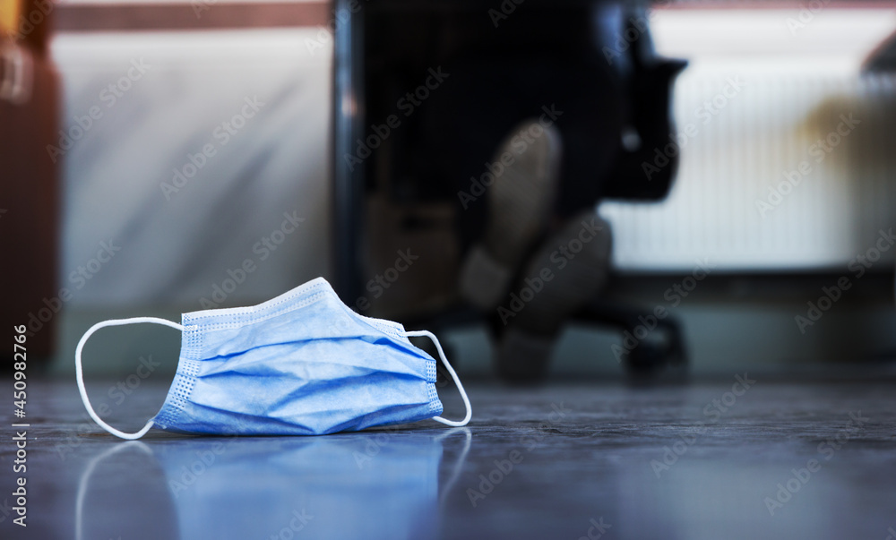 A medical mask abandoned on the floor of an office, with some working man's feet crossed over in the background. Concept for back to work after pandemic.