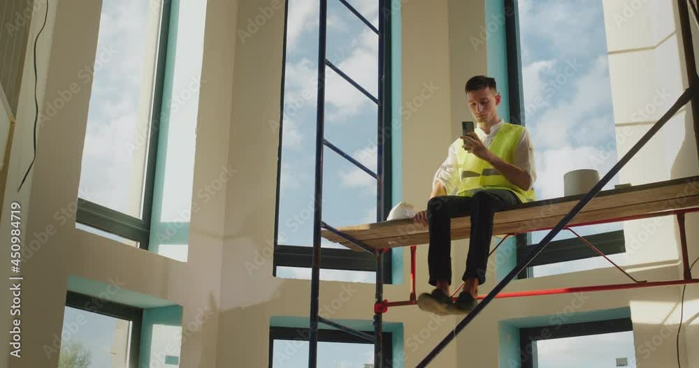 A bedless worker sits on scaffolding without a helmet, playing on a smartphone. Dangerous behavior concept A bedless worker sits on scaffolding without a helmet, playing on a smartphone. Dangerous behavior concept