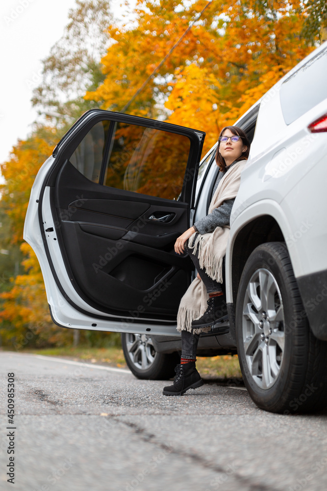 Handsome caucasian brunette woman in glasses, black boots, grey coat and beige scarf sitting in car doorway. One leg stay on asphalt. Girl enjoy autumn nature. Travelling by car. Selective focus.