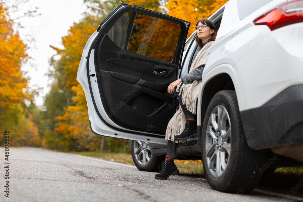 Attractive caucasian brunette woman in glasses, black boots, grey coat and beige scarf sitting in car doorway. One leg stay on asphalt. Girl delighting autumn nature. Travelling by car. Soft focus.