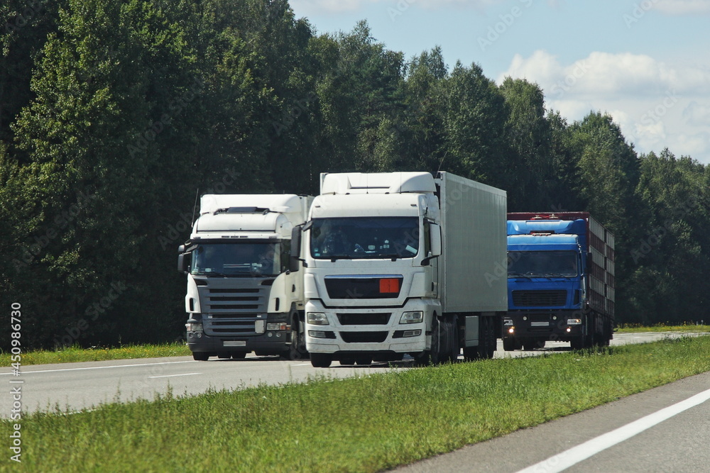 European semi trucks overtaking front side view into oncoming traffic drive on the suburban highway road at Sunny summer day on green forest and blue cloudy sky background, traffic landscape European semi trucks overtaking front side view into oncoming traffic drive on the suburban highway road at Sunny summer day on green forest and blue cloudy sky background, traffic landscape