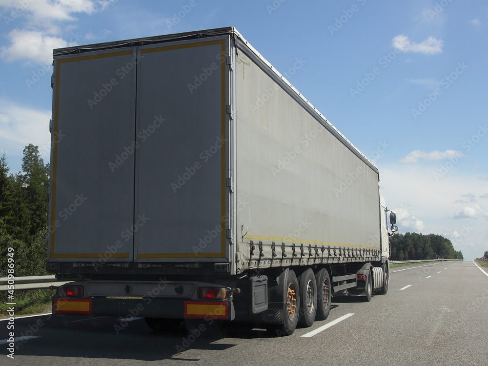 European heavy truck move on the countryside highway road at Sunny summer day on green forest, road fence and blue sky with clouds background, back side view, transportation logistics