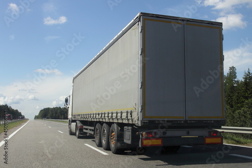 European heavy truck motion on traffic lane on the countryside highway road at Sunny summer day on blue sky and green forest background, back side view, goods delivery transportation logistics