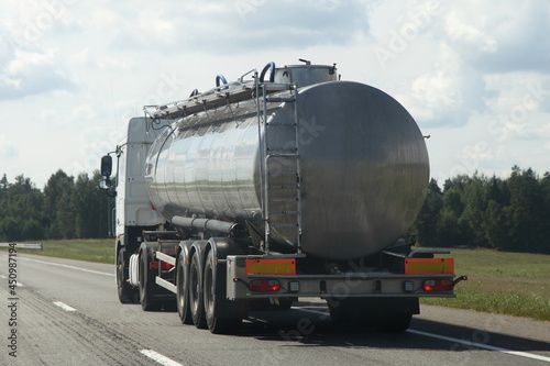 Gray barrel 3 axle big tank truck back side view on right lane move on the suburban highway road at Sunny summer day on blue cloudy sky background , liquid transportation logistics in Europe
