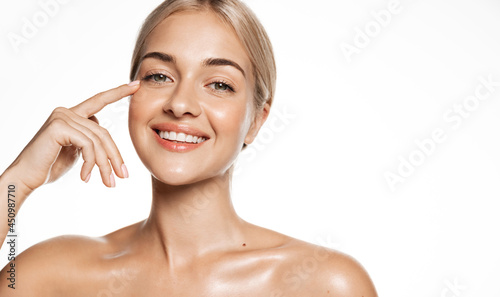 Young smiling woman with blond hair, gently touching facial skin with finger, concept of beauty spa and cosmetology. Girl without makeup with glowing skin after shower, head and shoulders portrait