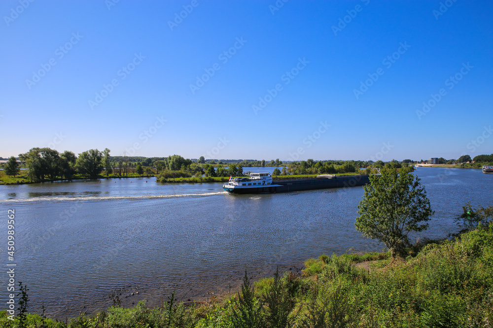 View over green rural landscape on river Maas with inland waterway vessel against blue summer sky - Between Roermond and Venlo, Netherlands