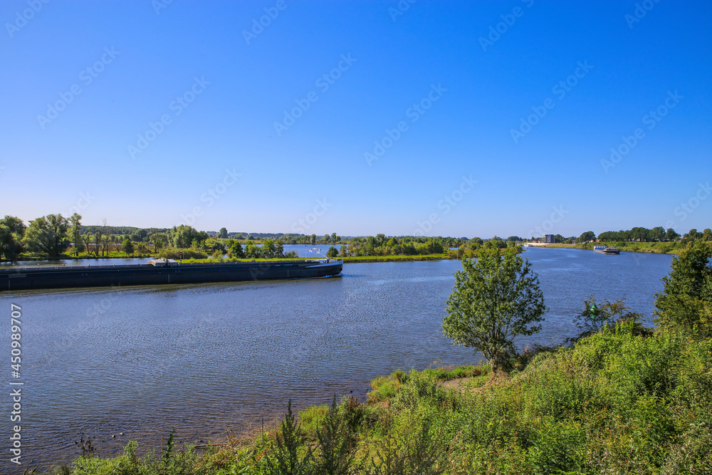 View over green rural landscape on river Maas with inland waterway vessel against blue summer sky - Between Roermond and Venlo, Netherlands