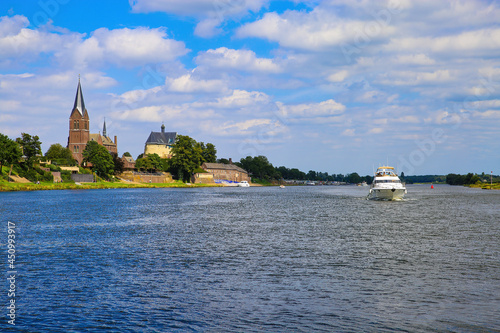 View over river maas on old villaga with church tower, motor boat against blue summer sky - Kessel, Netherlands photo