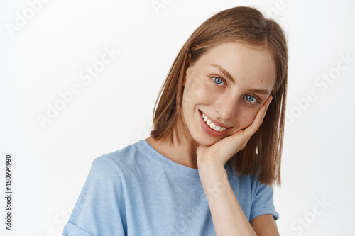 Women and beauty. Attractive young woman with fair short hair and smooth, glowing natural skin, touch her face and smiling white teeth, standing against white background © Cookie Studio