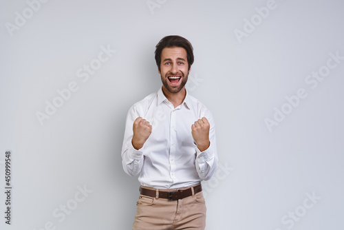 Happy young man in white shirt gesturing and looking at camera while standing against gray background
