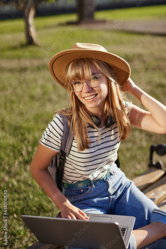 Homeschooling, e-learning, education or surfing concept. Attractive smiling blonde college or university student with backpack, eyeglasses and hat using laptop sitting on bench at university campus