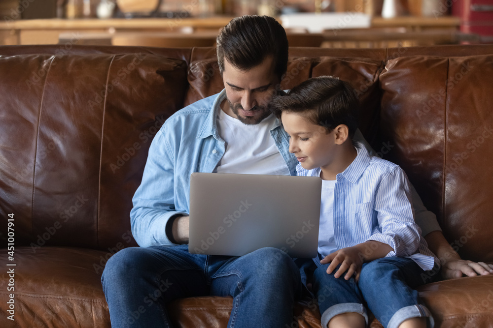 Young Caucasian dad and little teen son sit on couch in living room work together on laptop. Loving caring father have fun spend time with small teenage boy child use computer browse internet.