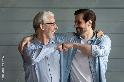 Overjoyed millennial man child and old Caucasian father isolate don grey wall background celebrate win or victory. Happy adult grownup son and mature dad give fist bump show unity and support.