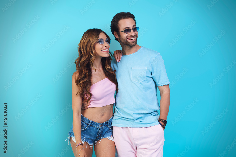 Beautiful young smiling couple embracing and looking away while standing against blue background Beautiful young smiling couple embracing and looking away while standing against blue background