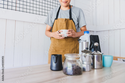 Smiling barista in brown apron is holding a cup of latte coffee in cafe shop