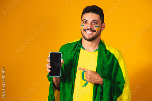 Brazilian fan. portrait, brazilian fan showing his cell phone, dressed as a football fan or soccer match on yellow background. Colors of Brazil. world Cup.