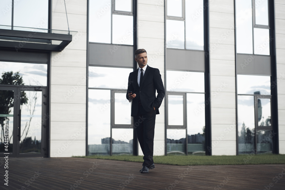Confident mature businessman holding mobile phone while walking in front of office building