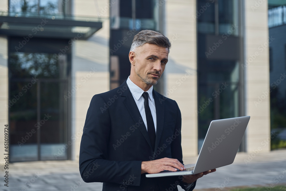 Confident mature businessman working on laptop while standing outdoors near office building