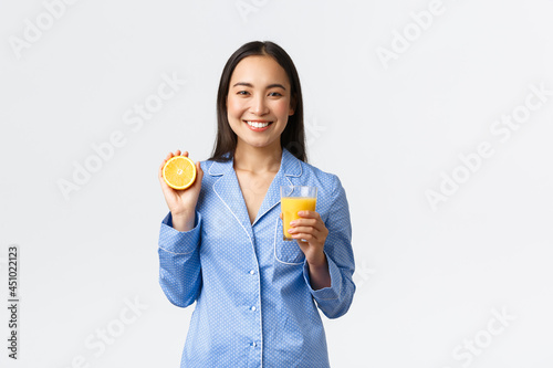 Morning, active and healthy lifestyle and home concept. Smiling cheerful asian girl starting her day with fresh made orange guice, holding glass and half of orange, looking happy and energized photo