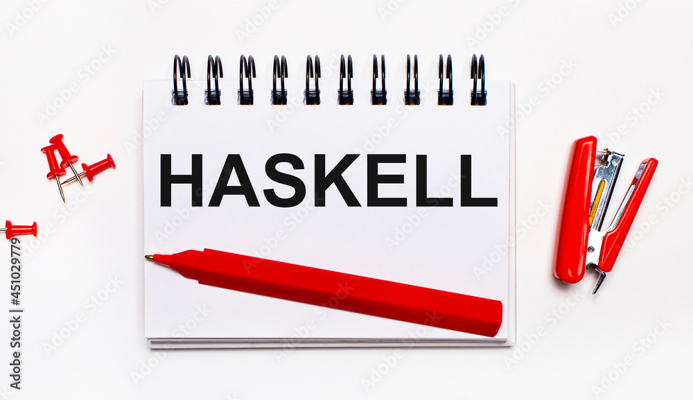 On a light background, a red pen, a red stapler, red paper clips and a notebook with the inscription HASKELL