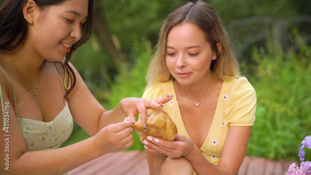 CU of asian and caucasian young woman eating fresh bred at the picnic. Female friends eating and talking, smiling. Young woman with Rheumatoid arthritis is eating bred CU of asian and caucasian young woman eating fresh bred at the picnic. Female friends eating and talking, smiling. Young woman with Rheumatoid arthritis is eating bred