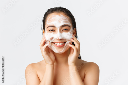 Skincare, women beauty, hygiene and personal care concept. Close-up of beautiful asian female standing naked in shower, apply skin cleansing foam and smiling, feeling fresh and upbeat photo