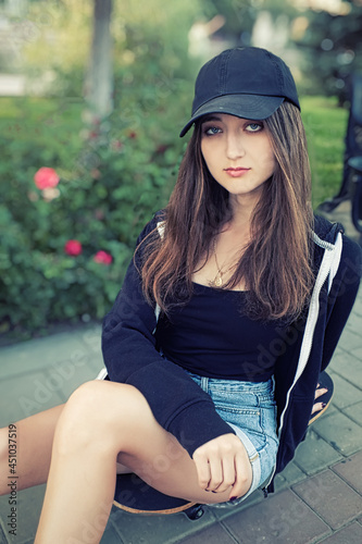 beautiful teenage lady with long legs in jeans short sits on skateboard in park setting. Her long silky hair on shoulders.