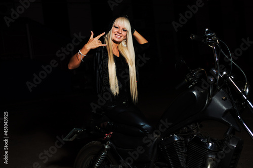 Blond haired biker girl is sitting on motorbike in night anf gesturing - shows mano cornuta or devils horns sign