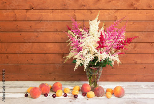 White, red and pink beautiful astilbe flowers in glass vase and fresh fruits on aged wooden table.